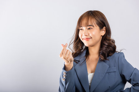 Confident business woman stands with a smile, creating a mini heart sign with her finger on a white background. Asian portrait of a beautiful young female sending love for Valentines Day, studio shotの写真素材