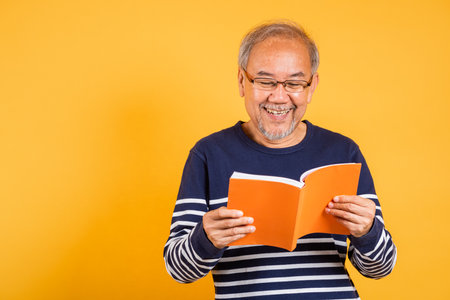 Portrait smiling Asian old man wearing glasses reading a book studio shot isolated yellow background, happy elderly man enjoying interesting story, time to relaxationの写真素材