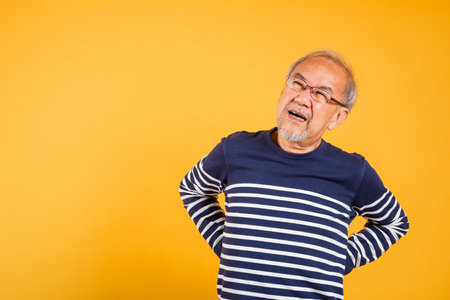 Portrait Asian old man with glasses holds hands on back he backache studio shot isolated yellow background, senior man pensioner suffering of backache, problems muscular painの写真素材