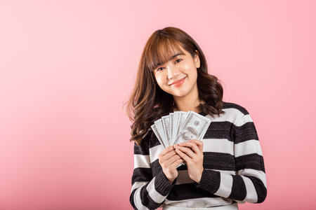 Asian happy portrait beautiful young woman smiling holding money fan banknotes 100 dollar bills isolated on pink backgroundの写真素材