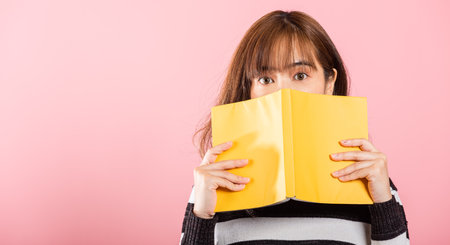 Portrait of beautiful Asian young woman teen smile covering her face with yellow book, female person...
