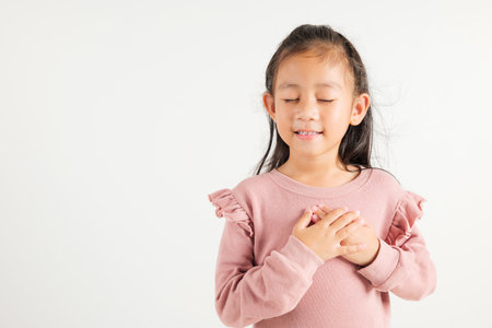 Happy young kid girl in casual clothes holding hands at chest close to heart meditation and smiling in studio short isolated on white background, kindergarten child believe faith and love gratitudeの写真素材