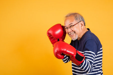 Portrait Asian old man wearing glasses wear two red boxing gloves studio shot isolated yellow background, smiling happy elderly man gray haired healthy fighter lifestyle conceptの写真素材
