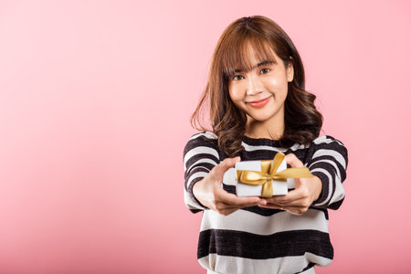 Portrait of happy beautiful Asian young woman smiling holding small gift box on hands, studio shot isolated on pink background, gift for you. Birthday, New year, Christmas, valentine day conceptの写真素材