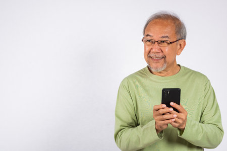 Portrait smiling Asian older man smiling holding a cell phone studio shot isolated on white background. elderly typing message on mobile phone in hand. Concept of connection and communicationの写真素材
