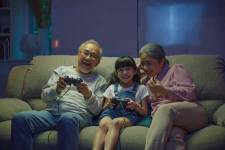 Happy Asian family bonding over a video game on a cozy couch, little girl playing video games console with senior grandparents together with controller joysticks while sitting on the sofa at nightの写真素材