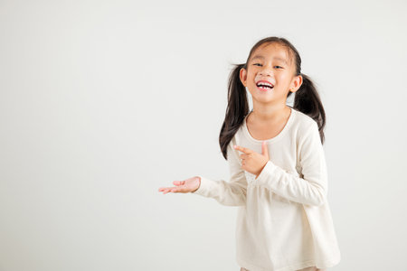 Asian happy portrait cute young girl smile presenting product holding something on palm away side, studio shot isolated on white background with copy space, kindergarten child show hand gestureの写真素材