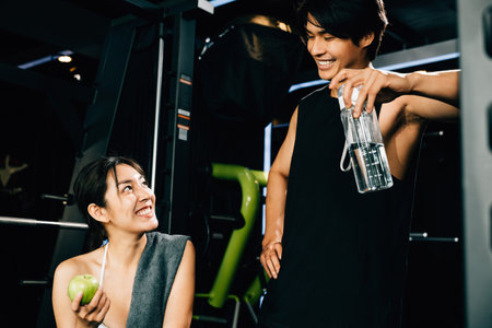 Young couple talking together after workout at gym, man hold water sport from bottle and woman holding green apple in fitness gym, eating healthy food after exercise, workout healthy lifestyle conceptの写真素材