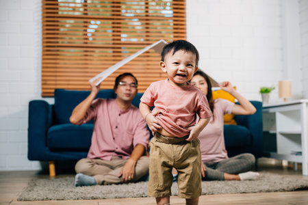 With their child a happy mother and father sit on the sofa holding a cardboard roof mockup symbolizing safety and protection during relocation at home. Happiness and family support is evident.の写真素材