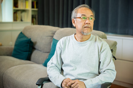Asian senior man disabled sitting alone on wheelchair in living room looking out window at home and thinking, lonely elder sad old man retirement memory loss or nostalgia thought, feel depressedの写真素材