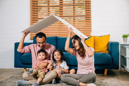 A smiling family sitting on a couch holds a cardboard roof symbolizing happiness and safety in their new home. Asian parents and their daughter radiate joy and togetherness.の写真素材