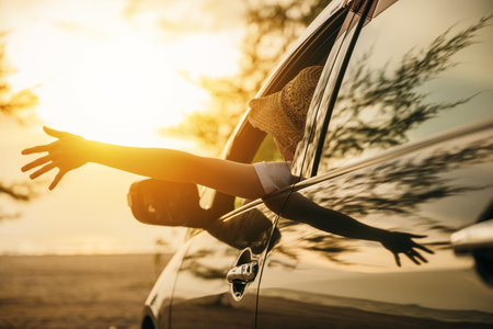 Woman wearing straw hat sits in front seat waving hand out car window at sunset feeling the wind. Happy weekend travel enjoy summer at beach. summer vacation travel driving road trip conceptの写真素材
