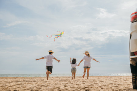 Asian family is flying a kite while running on the beach. Happy parents mother and father with their child playing with kite, The car is parked nearby, and the family is enjoying their time togetherの写真素材
