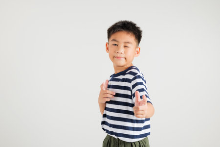 Cheerful Asian young kid boy excitedly makes two gun finger gesture to the camera with a friendly and cheeky expression. Isolated on white background, primary child exudes positivity and joyの写真素材
