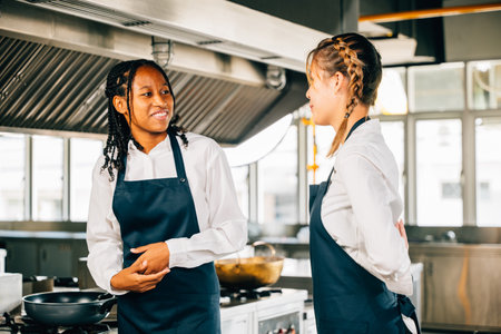 In a restaurant kitchen a girlfriend chef assists her friend in tying an apron. Two women in uniform prepare for food education and service.の写真素材