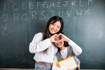 Two women are holding hands and making a heart shape on a chalkboard. They are smiling and seem to...