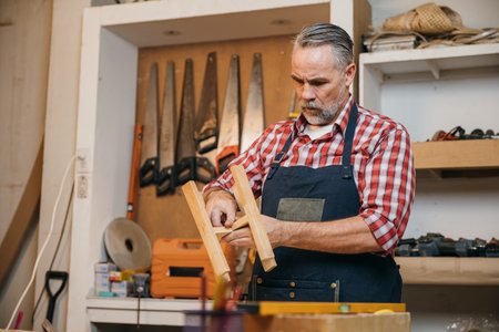 Senior carpenter in a plaid shirt and apron assembling a wooden stool in a workshop. woodworking skills, carpentry, and hands-on craftsmanship in a workshop setting, National Carpenters Dayの写真素材