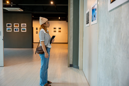 Asian woman holding tablet at art gallery collection in front framed paintings pictures on white wall, Young person at photo frame hold digital book leaning against at show exhibition artwork galleryの写真素材