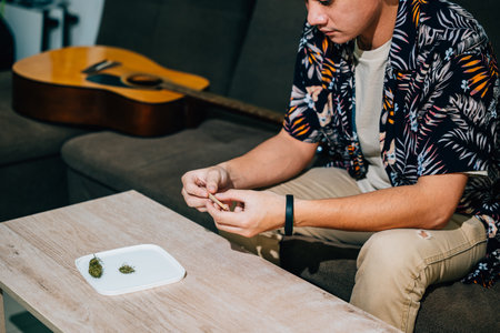 Asian young man sitting on a couch with a cannabis bud in his hand and smoking a joint relaxing time at home. A guitar is in the backgroundの写真素材