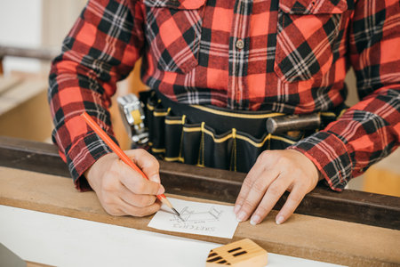 Person in a red plaid shirt sketching table plans on paper with a red pencil, surrounded by wooden...
