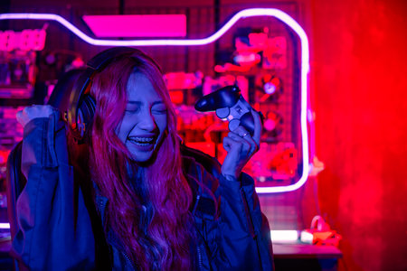 Winning. Happy Gamer using joystick controller for virtual tournament plays online video game with computer she raises hands to wins celebrating, Asian woman wearing gaming headphones feeling excitedの写真素材