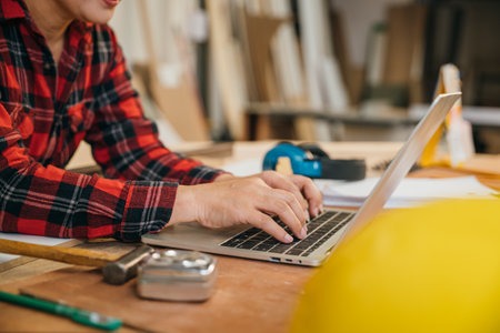 Carpenter woman in a white hardhat and red plaid shirt uses a laptop in a woodshop, surrounded by tools, wood scraps, and pencils. Ideal for online learning, client orders, or woodworking projectsの写真素材