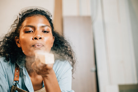 African American carpenter woman with curly hair and safety glasses blowing sawdust off wood in DIY workshop. Empowering female joiner confidently working in carpentry industry, National Carpentersの写真素材