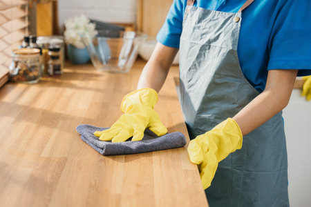 Maid in protective glove cleans wood table in cozy kitchen. Using professional cleaning products for home perfect tidiness. Cleaner working safety glove hygiene routine. Maid housekeeping concept.の写真素材