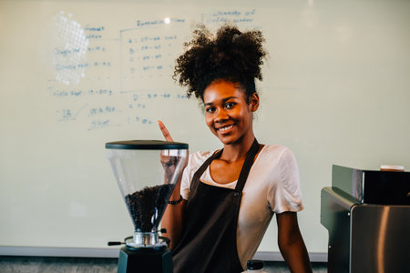 In the coffee shop confident black woman stands at the counter. Portrait of owner a successful businesswoman in uniform smiling with satisfaction providing great service. Inside a small business cafeの写真素材