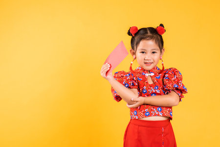 Chinese New Year. Happy Asian Chinese little girl smile wear red cheongsam qipao hold angpao red packet monetary gift, Portrait child in traditional dress, studio short isolated on yellow backgroundの写真素材