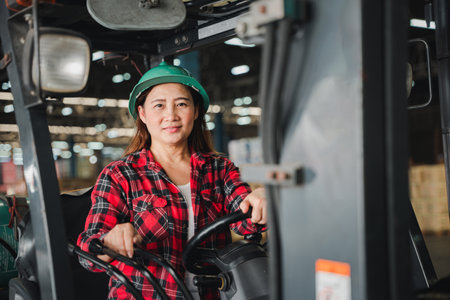 At the factory an Asian woman engineer skillfully drives a forklift truck indoors portraying an adult female professional in an industrial setting managing tasks within the warehouseの写真素材