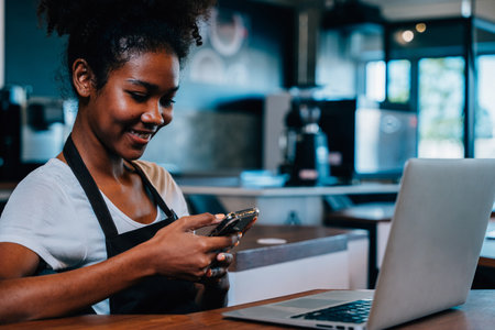 Portrait of cafe owner a concentrated black woman in apron using smartphone in her shop. Multi tasking barista manages orders communicates runs her cafe ensuring excellent service.の写真素材