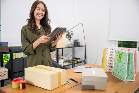 Asian smile young woman checking online order packing box on digital tablet before shipping to customer at home office, startup small business on websites and social mediaの写真素材