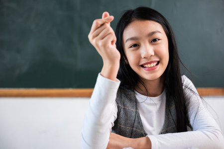 A young girl is smiling and holding her hand up in front of a blackboard. She is wearing a white shirt and a gray vestの写真素材