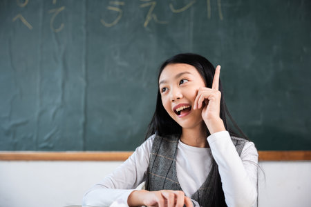 A girl is sitting in a classroom and pointing her finger. She is smiling and looking at the chalkboardの写真素材