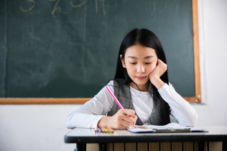 A girl is sitting at a desk and writing with a pink pencil. She is wearing a white shirt and a gray vest. The room is decorated with a blackboard and a chalkboardの写真素材