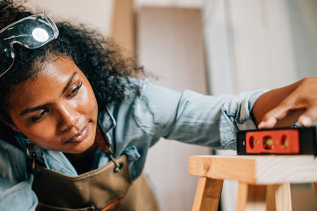 African American carpenter woman uses a level on a wooden stool in the workshop. Master joiner skillfully crafting solid pine furniture, surrounded by tools and materials, National Carpenters Dayの写真素材