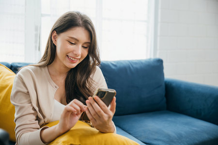 A smiling woman casually sitting on a sofa holds her phone typing messages. Amid home comfort she finds success in communication and technology radiating relaxation. The woman is relaxing.の写真素材