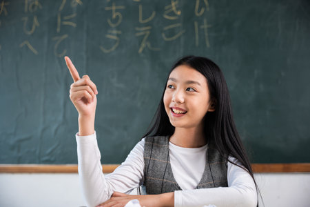 A young girl is sitting at a desk in a classroom with a green chalkboard behind her. She is pointing to the board with her finger, possibly indicating something important or interestingの写真素材