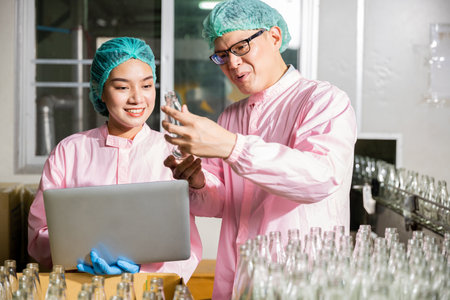 Inspectors scrutinize product bottles moving on a beverage factorys conveyor belt. Quality control and inspection managed by engineers using a laptop for precise analysis.の写真素材