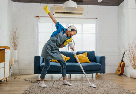 House cleaning becomes delightful Asian housewife joyfully sings using a vacuum cleaner as a microphone during cleanup. Dancing and cleaning in living room a cheerful maid enjoying her work.の写真素材