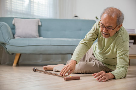 Sick senior old man falling down lying on floor because stumbled at home alone with wooden cane walking stick in living room, elderly man retired grandfather having accident after standing from sofaの写真素材