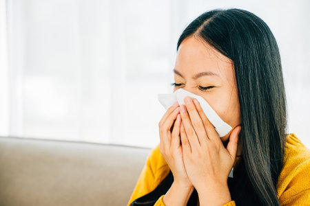 A woman at home holding a tissue experiencing symptoms like coughing and stuffy nose. Portrait of a patient dealing with illness. Snot and handkerchief in hand.の写真素材