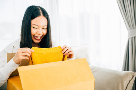 Sitting on a couch an Asian woman unboxes a delivered dress from online shopping. Her smiling face exhibits happiness and contentment with the received item. Satisfied buyer concept.の写真素材