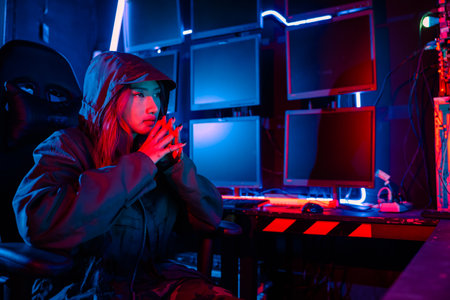 Woman hacker. A woman wearing a hood is thinking looking at a computer monitor. The room is dimly lit. possibly typing or browsing the internet, Hacker in neon light, Female gamer thinkの写真素材