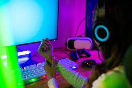 Young Asian woman gamer wearing headphones playing video game on modern gaming pc computer have long time she wrist pain, female broadcast streaming plays online home games holding wrist her problemの写真素材