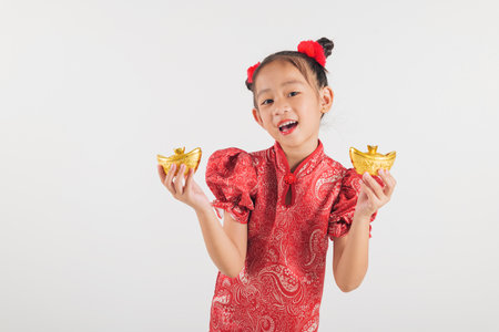 Festive Asian child in a red cheongsam dress holding a gold ingot. Isolated on a white background, celebrating Chinese New Year traditions and spreading joy and prosperity with her bright smile.の写真素材