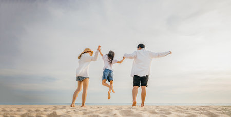 A moment of pure joy on beach as an Asian family father and mother holding children gleefully jump into air. beautiful ocean provides serene background to this delightful expression of family fun.の写真素材