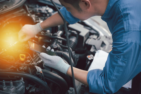 Smiling technician taking a break at car workshop, texting on mobile phone. Close-up shot of mechanics hand holding wrench while standing near car engine. Horizontal photo with workshop background.の写真素材