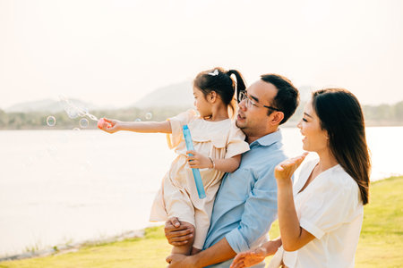 Asian family having fun playing together on spring meadow field grass in park, Happy parents father carrying daughter and a child blowing soap bubbles enjoying outdoors together with motherの写真素材